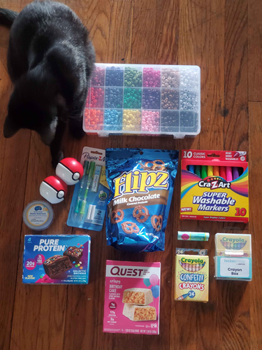 A photo of miscellaneous birthday gifts laid out neatly on the floor. A black kitten in the top left bats at some of the gifts curiously.