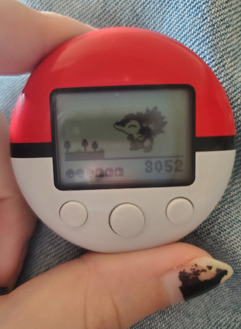 An image of a Pokewalker, a red and white circular pedometer in the shape of a Pokeball. The screen shows a Cyndaquil with 3,052 steps, three items found, and two captures.